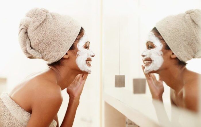 Mirror reflection of a young ethnic woman laughing and applying a face mask to her face