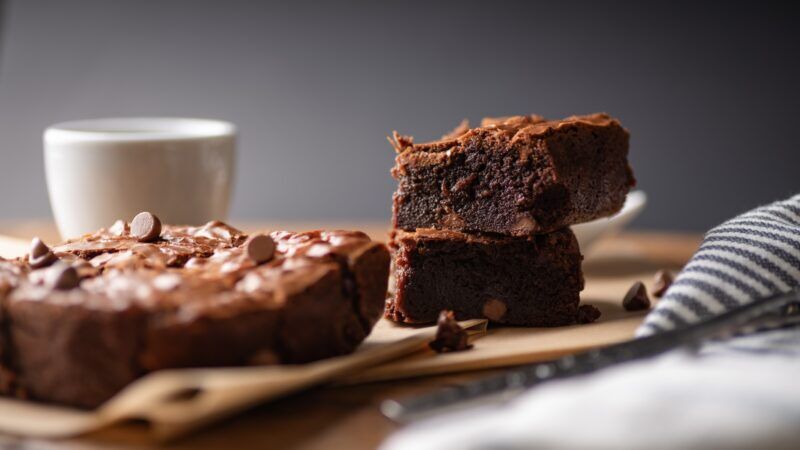 Chocolate chip brownies accompanied by coffee and chocolate chips, presented on a wooden board alongside a fork and knife, with baking paper enhancing
